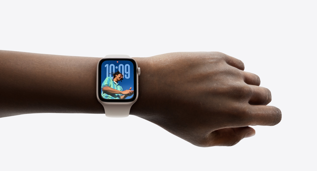 Lähikuva hopeanvärisestä Apple Watchista ja silikonikumirannekkeesta ranteessa, näkyvissä Aika-appi uudella Liquid Glass ‑ulkoasulla