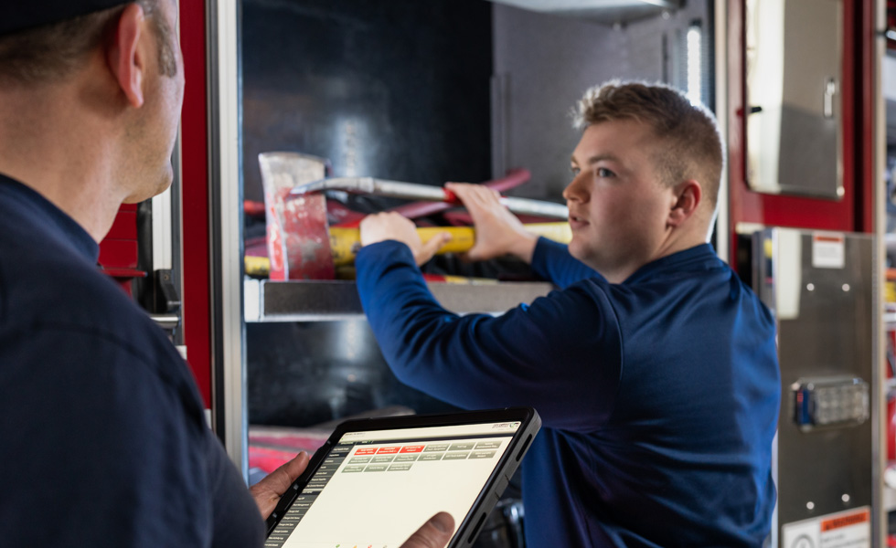 Zwei Feuerwehrmänner überprüfen Werkzeuge und Ausrüstung in einem Feuerwehrauto, einer hält ein iPad, während der andere kontrolliert 