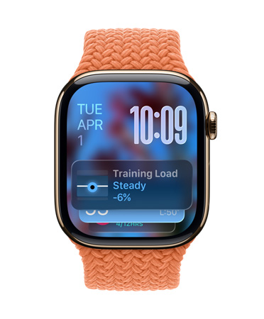 Ruusukullan värinen Apple Watch ja punottu mandariininoranssi ranneke, kellotaulun vasemmassa yläkulmassa näkyy päivämäärä, oikeassa yläkulmassa näkyvissä numerot päällekkäin, Treenikuorma-widgetti alhaalla, kaikki läpikuultavalla Liquid Glass ‑käyttöliittymällä